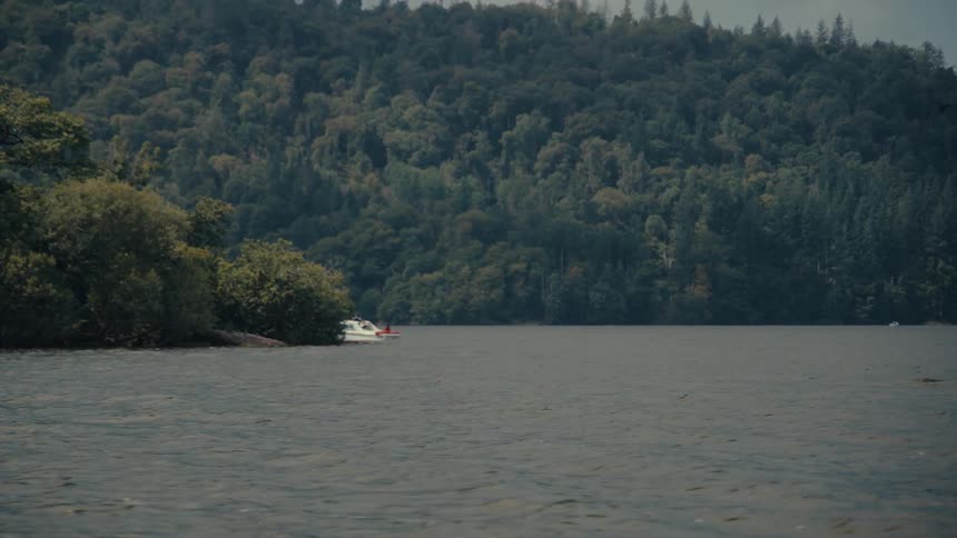 A small white boat moves slowly across a wide lake surrounded by a dense forested shoreline the choppy water and overcast sky give the scene a serene slightly moody atmosphere