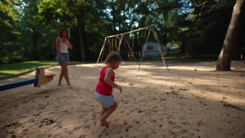 A child with light hair and a red top walks across a sandy playground toward the left while a woman in a white tank top stands in the background near a seesaw and a set of swings