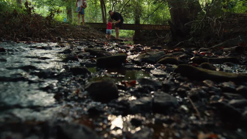 The camera low to the ground shows a shallow stream of water flowing over smooth dark rocks in the background three people stand on a small wooden bridge enjoying a sunny day in the woods