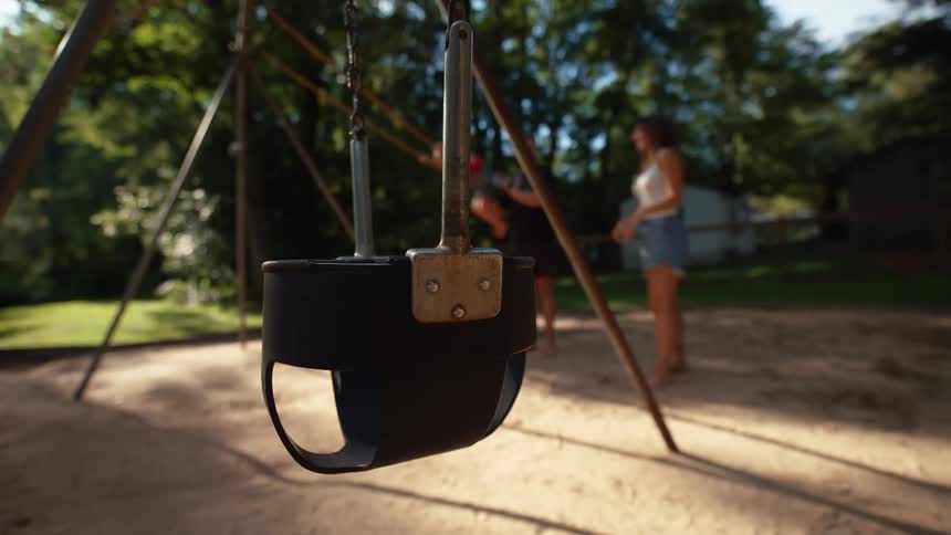 A dark infant swing hangs in the foreground with a family playing on a swing set in a sandy park behind it