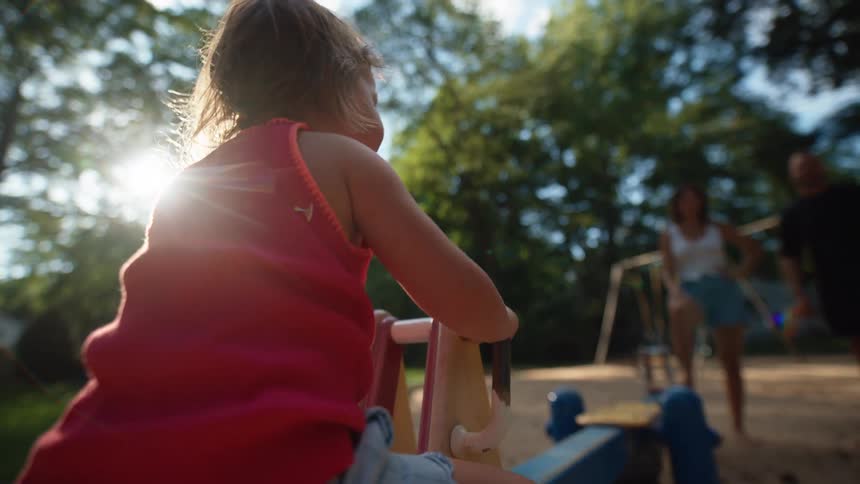 A little girl backlit by the sun plays on a rocking horse at a sunny park in the background two people stand on the sandy ground chatting and enjoying the day