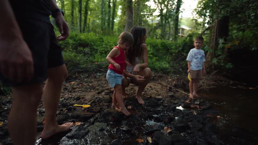 A father and mother and their two children are playing together in a small stream in a forest where they are all standing barefoot on the rocks and digging in the mud
