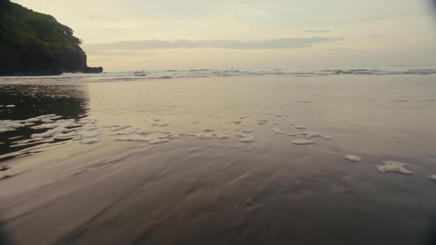 A shot focuses on a misty rocky coastline at dusk with gentle waves rolling in under a pale overcast sky the camera is positioned low capturing the foam of the waves as they wash onto the dark sand