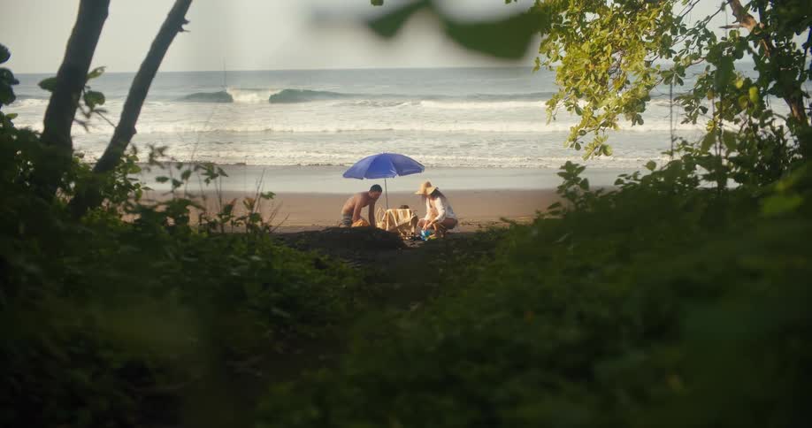 The video shows two people sitting on a beach under a blue umbrella with the ocean waves crashing behind them they appear to be enjoying a picnic