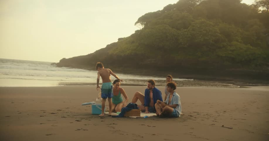 A family is having a beachside picnic on a large expanse of sand as the sun begins to set two children run and play while the parents relax and talk on a picnic blanket nearby