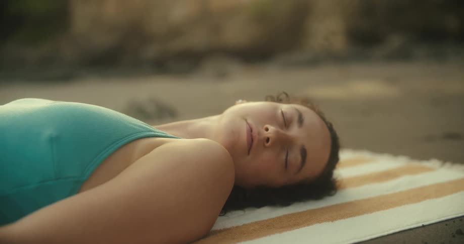 A woman with dark hair and a light blue top is lying on a striped towel on the ground with her eyes closed