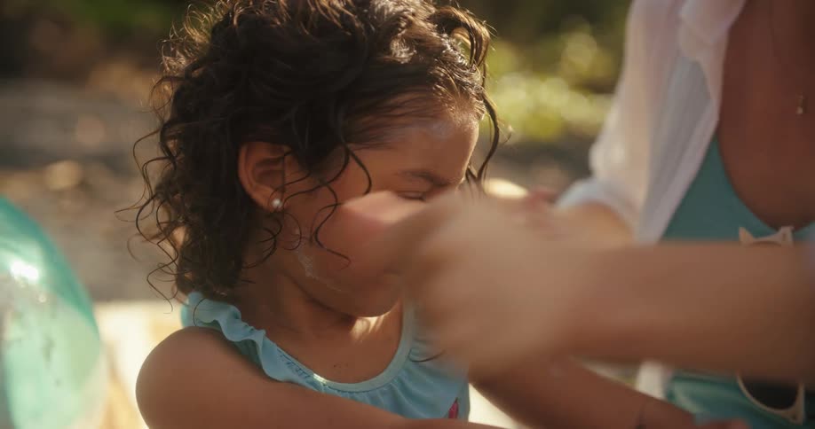 A mother carefully applies sunscreen to her young daughters back while the child stands patiently the child has wet curly hair and a focused expression as her mothers hands move across her skin