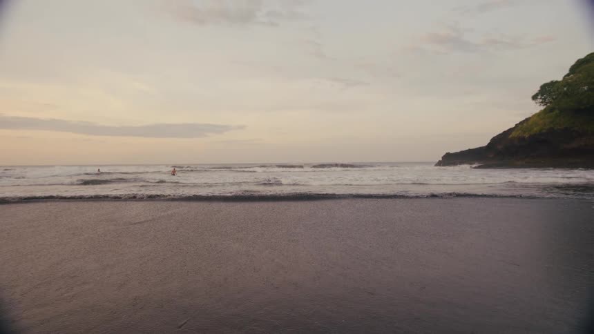 A lowangle shot captures the gentle waves of the ocean rolling onto a dark sand beach at dusk the tide washes in around a small rock with a large green hillside in the background