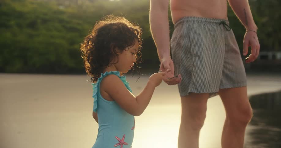 A person and a child stand on a beach then the child lets go of the persons hand and runs toward the water