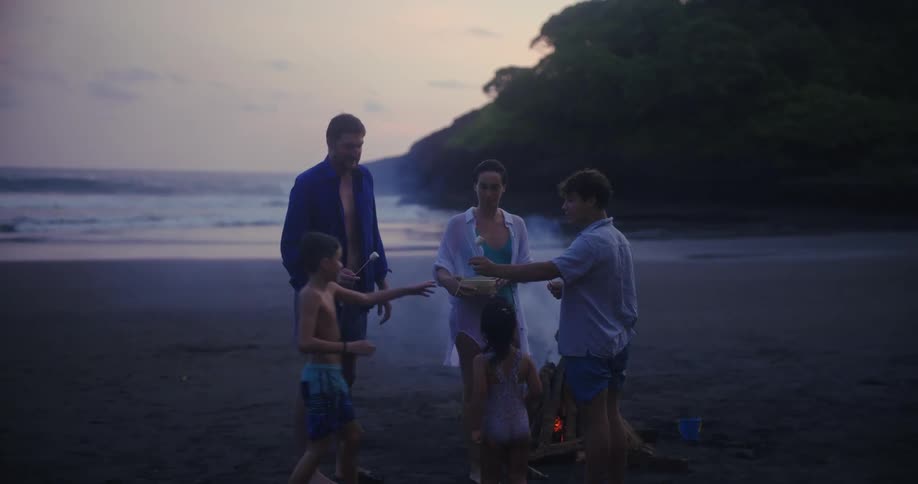 A family of four gathers around a small bonfire on a dark sand beach at dusk preparing to roast marshmallows the waves crash gently in the background as the parents pass out sticks to their excited children