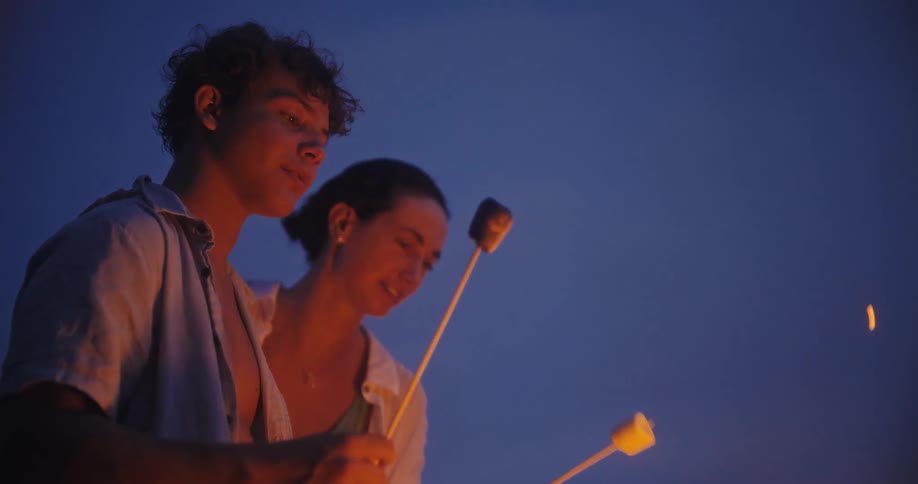 Young people roast marshmallows on sticks at night with the flame lighting up their faces