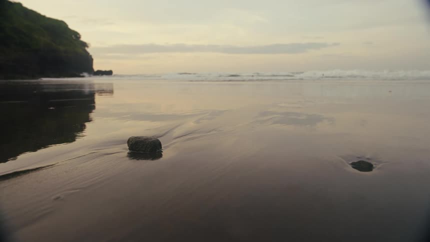 A lowangle shot captures the gentle waves of the ocean washing onto a dark sand beach at dusk the tide recedes around a solitary rock creating a fleeting trail in the wet sand