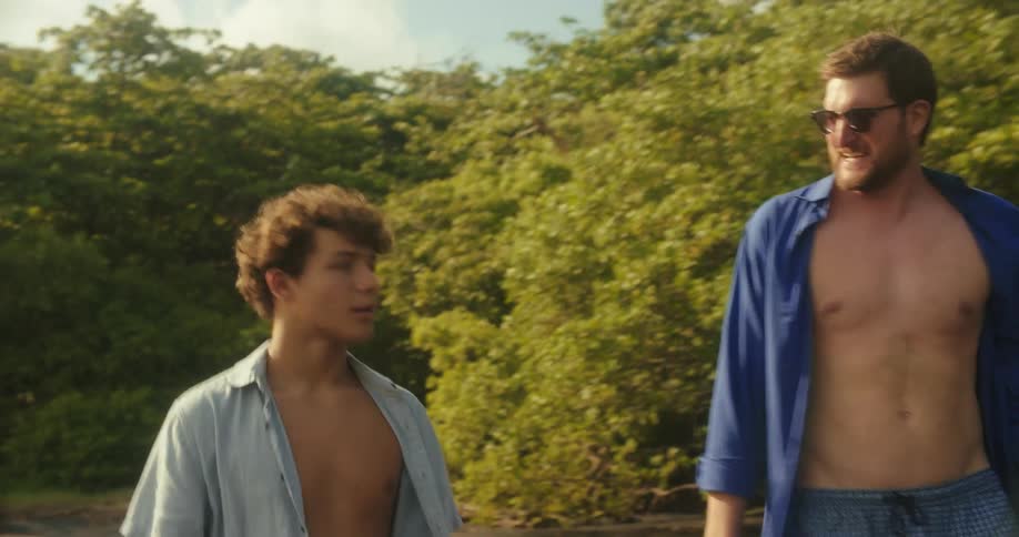 The video shows two men walking together on what appears to be a beach or a shoreline they are both shirtless and the older man in the blue shirt and sunglasses points to something in the distance