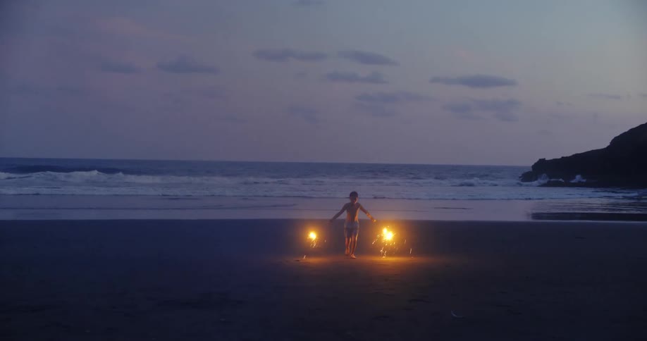 Young boy running with sparklers on a sandy beach at dusk