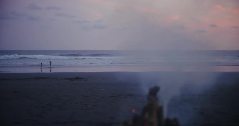 Two people walk along the seashore at dusk with smoke from a bonfire in the foreground