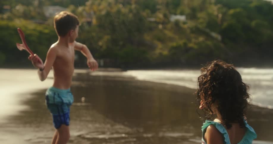 A boy on a beach holds a boomerang while a girl stands nearby