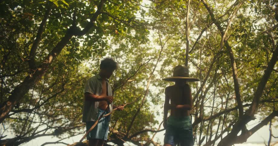 The video shows two young men on a beach in a lush tropical environment one of the men is wearing a hat and is bent over looking at something in the sand while the other is standing and watching him the scene is bright and has a warm adventurous fee