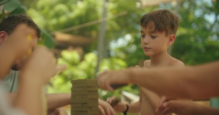 People sit at a table outdoors and play with a tower of wooden blocks