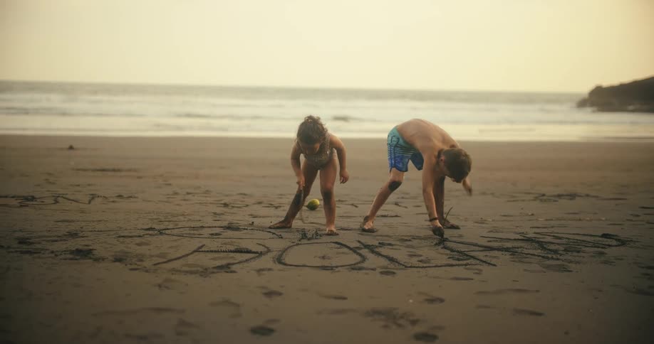 A young girl and boy are happily drawing in the sand on a beach at sunset