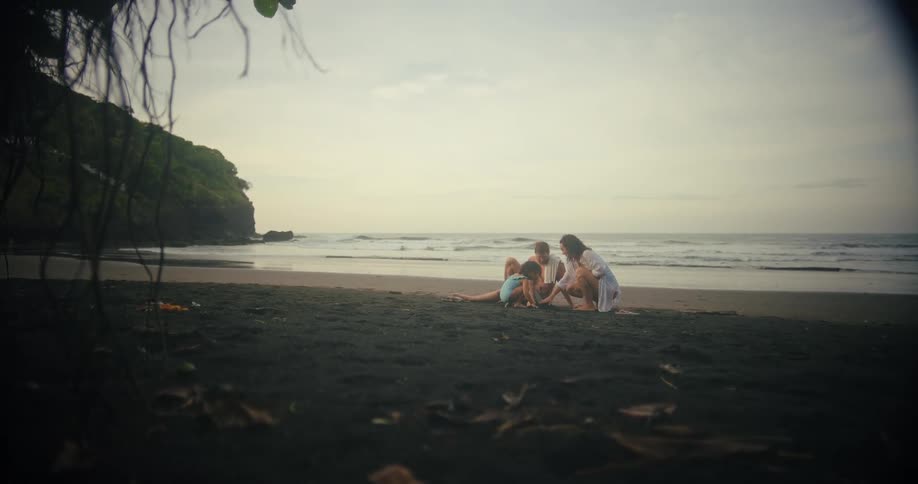 A family sit on a dark sand beach at the edge of the water as waves break on the shore with a large rock formation covered with plants