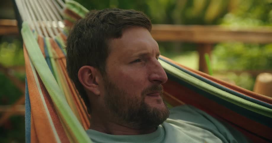 A bearded man with a content expression relaxes in a colorful striped hammock gently swaying in a peaceful outdoor setting