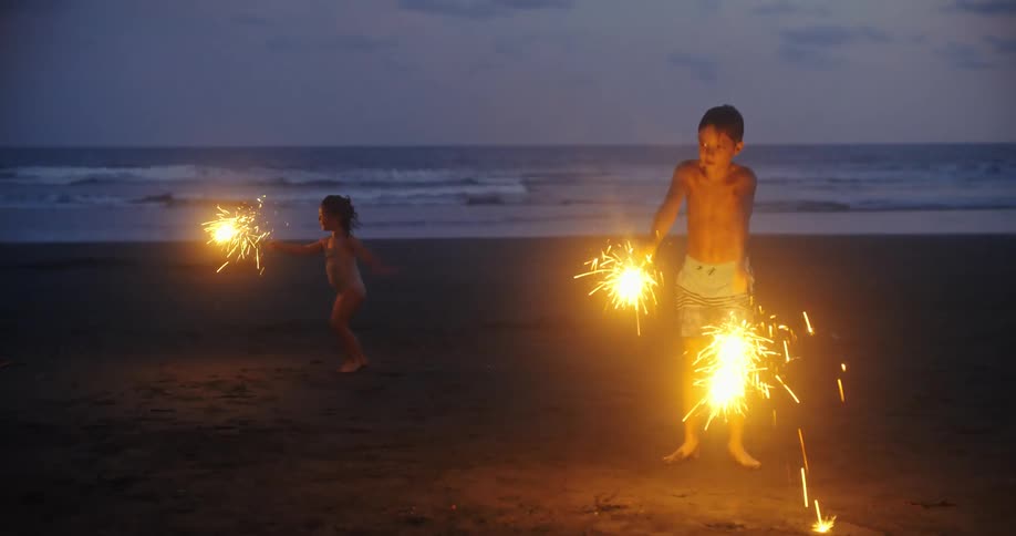 A young boy and girl are playing with sparklers at night on the beach