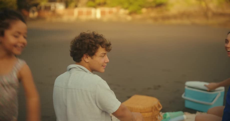 Two children run on a beach and a person holds a towel over their head while the other person follows then they meet their family sit on the sand