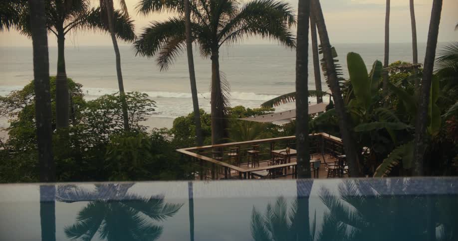 A patio with tables and chairs located near the ocean is surrounded by palm trees