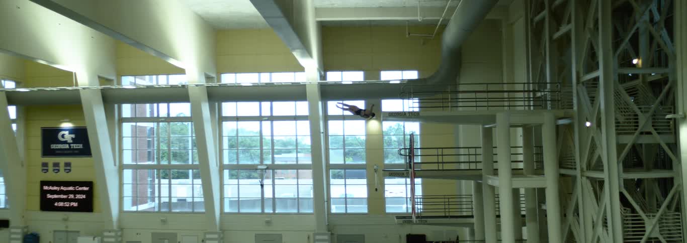 A man is captured in midair after jumping from a high diving board he is positioned horizontally with his body parallel to the water below and is wearing a black swimsuit the shot is taken inside with large windows visible in the background