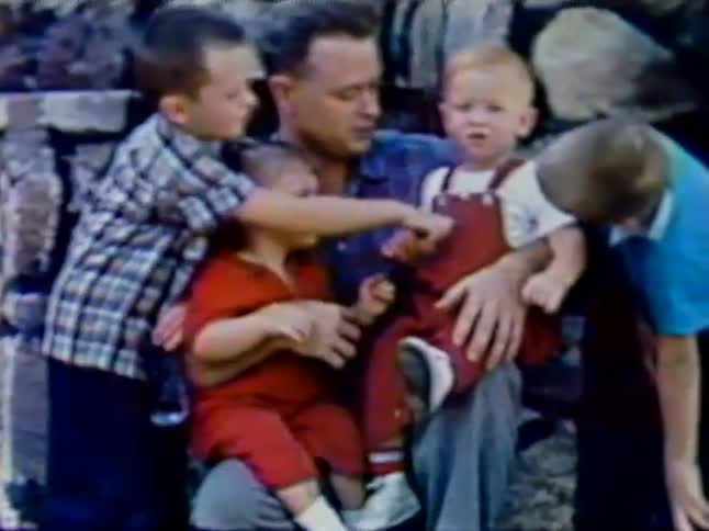 A man holds two children on his lap as two stand by him