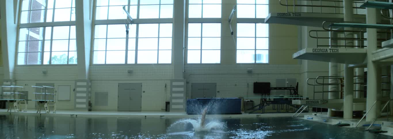 A diver in a dark swimsuit is shown mid air after jumping from the highest diving platform gracefully entering the pool and making a small splash