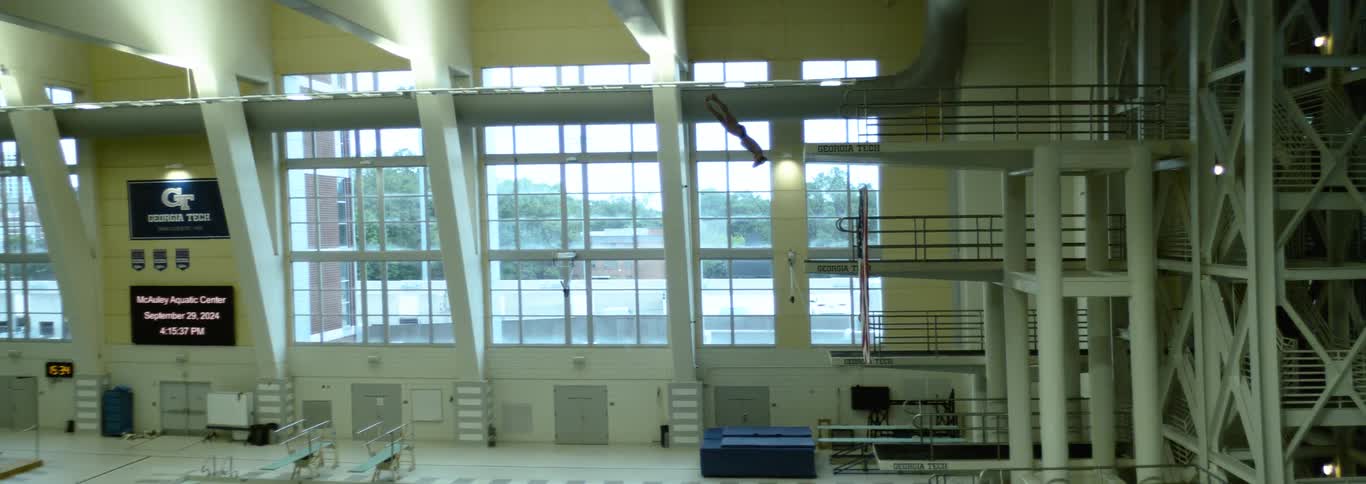 A person jumps from a high diving board turns in the air and falls toward a swimming pool with bleachers on the side