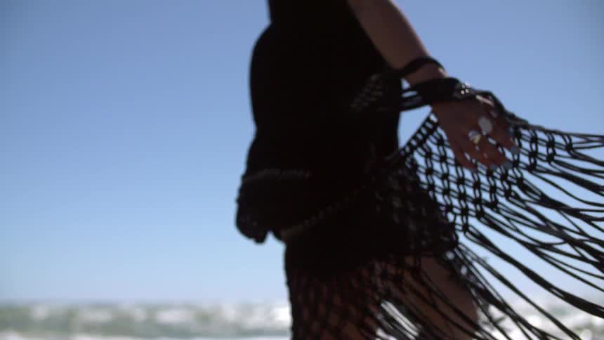 A woman wearing a black dress and shawl twirling on a beach as the sun reflects off waves