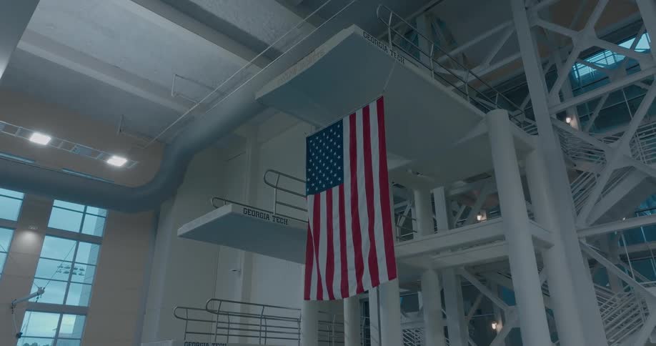 A close view of a tall white diving platform shows multiple diving boards each with the words georgia tech on them with a large american flag hanging down between the platforms