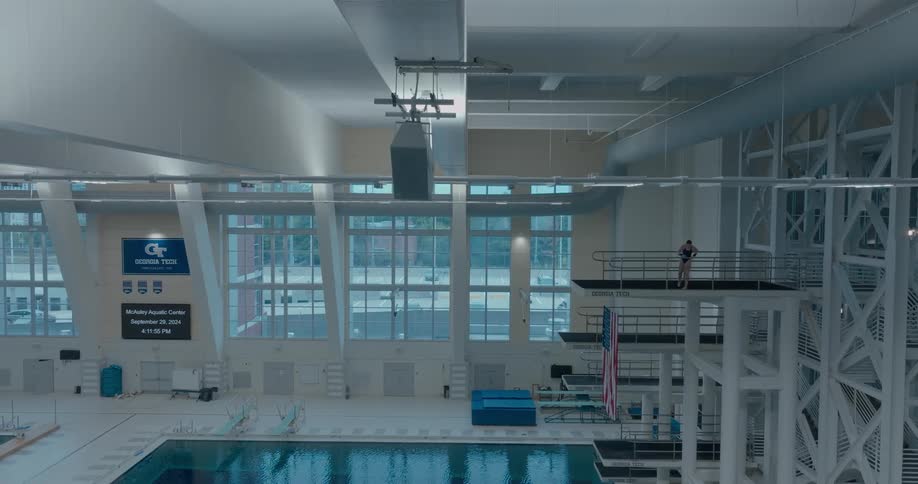 A wide view shows a female diver standing on a high platform in an indoor swimming facility that has large windows on the far wall