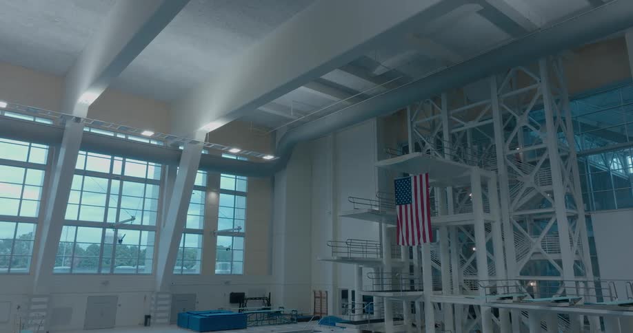 A high ceiling with metal rafters hangs over a large indoor pool facility which features a tall diving platform with a flag hanging from it and large windows on the far left