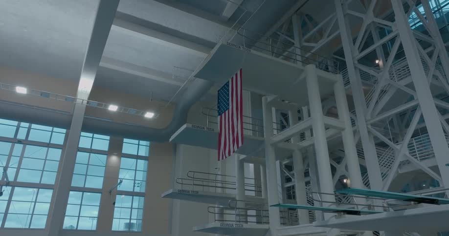 The video shows an american flag hanging in a large indoor space it is attached to a high ceiling and a large window is visible in the background suggesting the space is a gymnasium or similar large building