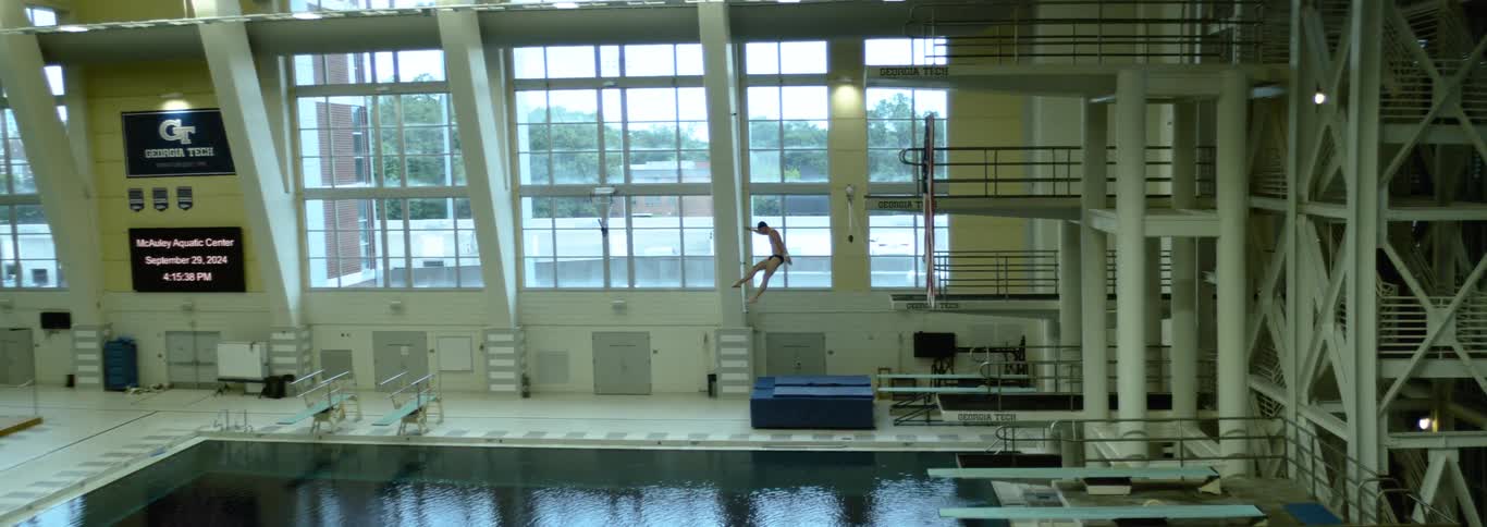 A professional diver is captured midair during a dive with their body fully extended over the water the dive takes place in an indoor aquatic center with large windows in the background revealing a view of trees outside