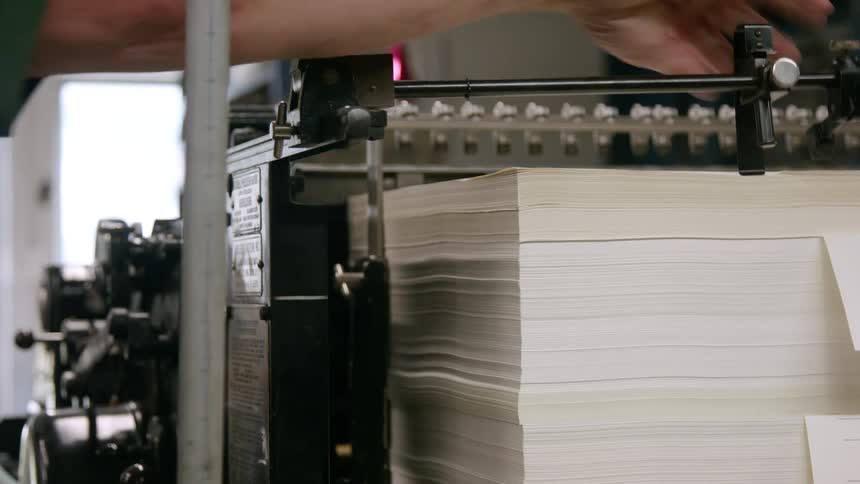 The camera shows a closeup of a machine moving a large stack of paper while a mans hand adjusts the machine