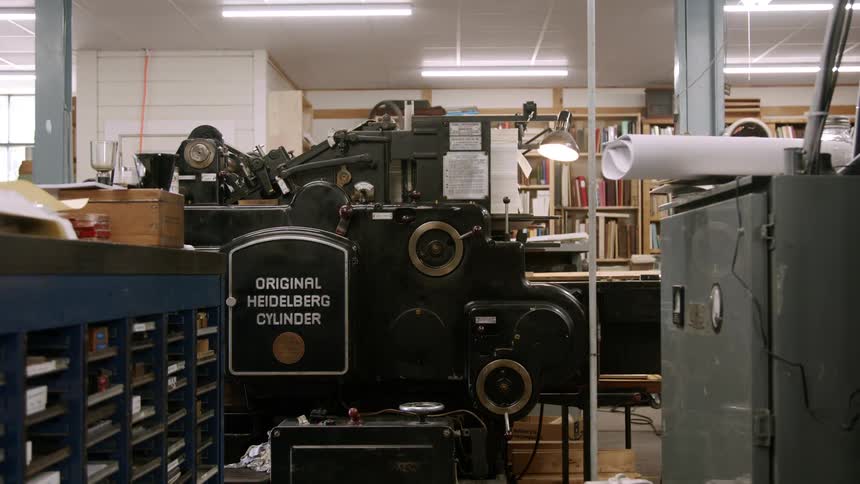 A wide shot of a printing workshop shows a large vintagelooking original heidelberg cylinder press in the center of the room the room is cluttered with various tools paper and stacks of books giving it a livedin creative feel