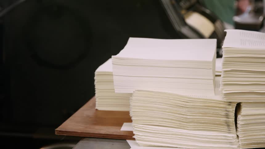 Freshly cut stacks of paper sit beside a running press in a print shop