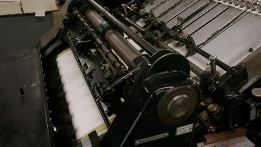 A wide view of a large dark printing press machine shows a large roll of brown paper feeding into the machine while white paper with black text is pushed out on a flat surface above