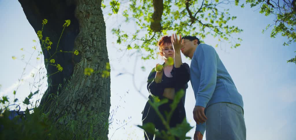 A young man and a young woman stand beside a tree at midday with the woman making hand movements as the young man watches her