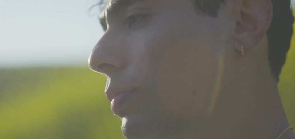 A young man with a small earring looks thoughtfully into the distance a slight smile playing on his lips as sunlight creates a soft lens flare