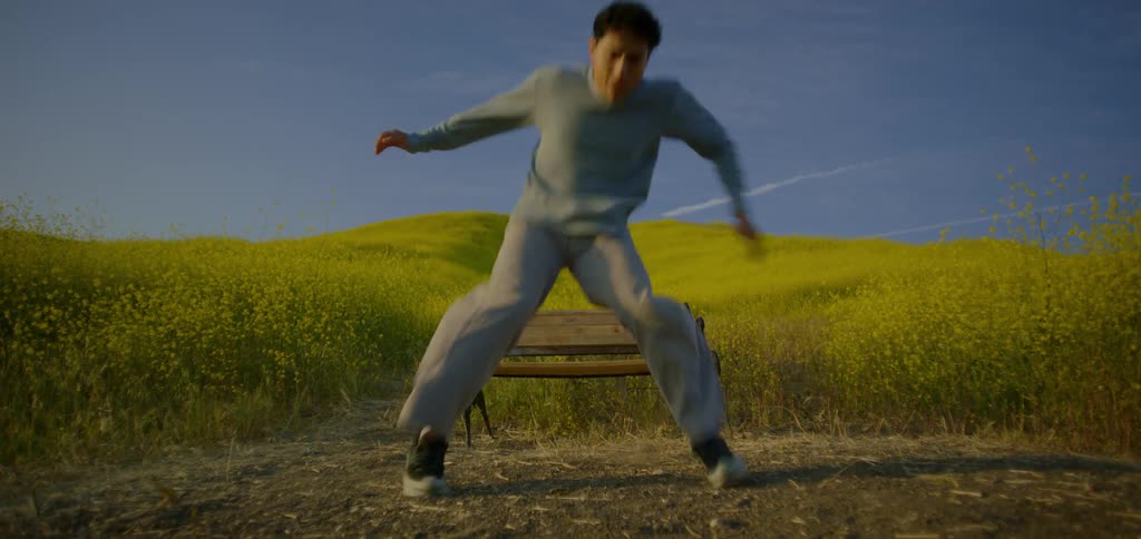 A shot focuses on a man dancing and kicking in a field of bright yellow flowers the man is jumping and kicking with a bench in the background of the shot