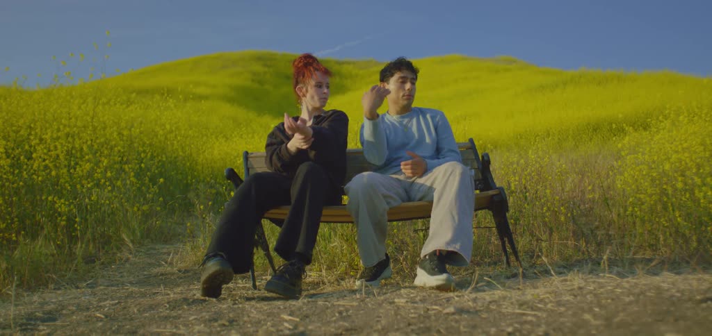 A shot focuses on a man and a woman sitting on a bench in a field of yellow flowers the man is seen looking ahead while the woman looks at him as she smiles
