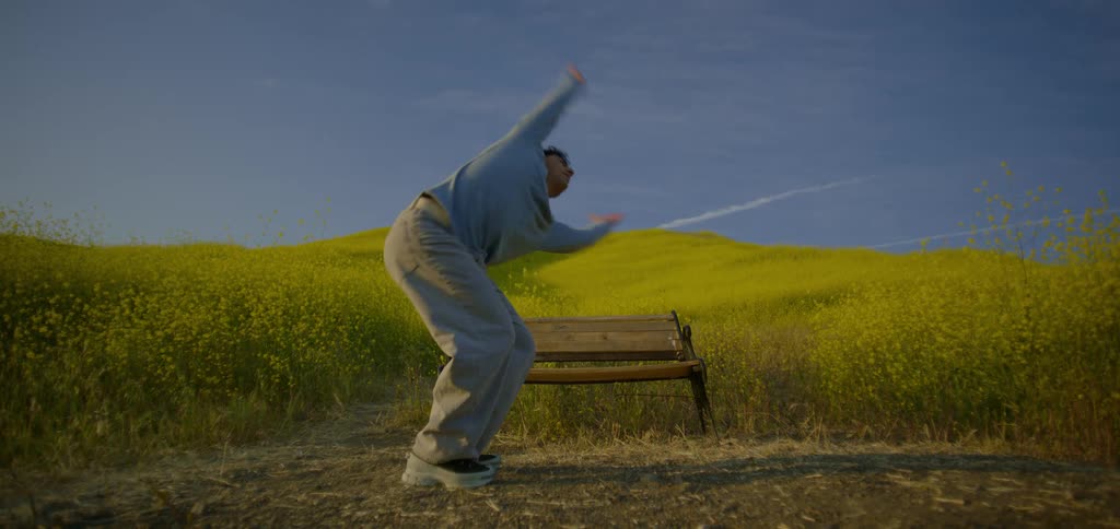 A man wearing a grey sweater and gray pants is dancing with expressive movements in front of a park bench in a wide field of yellow flowers under a blue sky