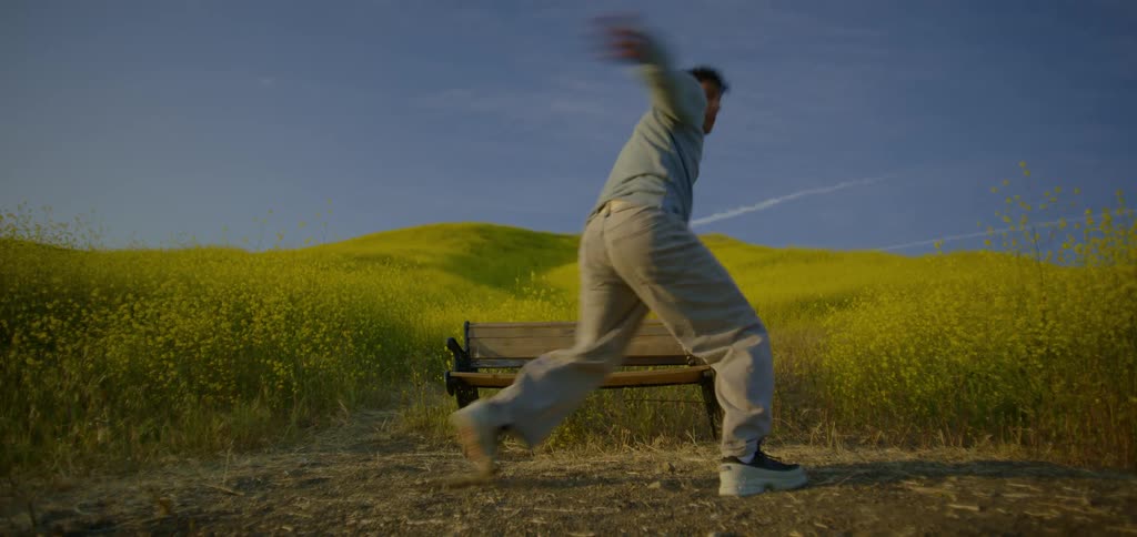 A shot from the side shows a person wearing a gray sweater and gray pants as they stumble and flail their arms the person is standing in a field of yellow flowers and green grass with a wooden bench behind them