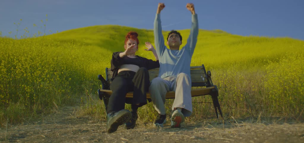 Two dancers sit on a park bench in a vibrant yellow field striking a pose the woman has red hair and is wearing all black while the man is in a light blue shirt and gray pants