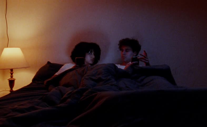 Two people with dark curly hair are lying in bed using their phones a warm dim light from a lamp illuminates the scene as they scroll through their devices